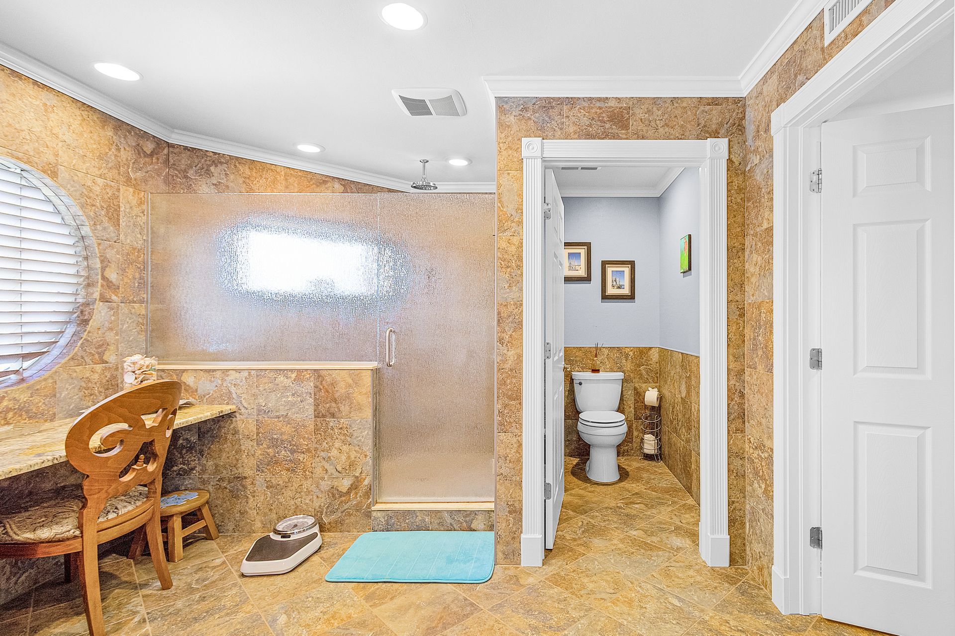 Small tiled room with a toilet, desk, chair, and shower, lit by bright overhead lights