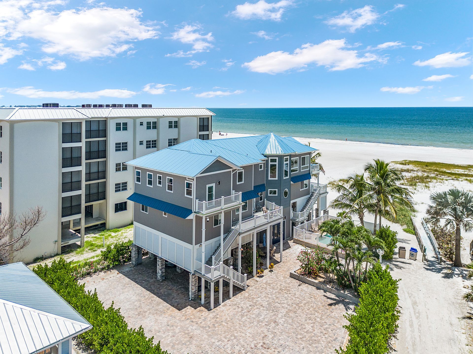Blue beach house on stilts beside a white condo, with palm trees and ocean under a sunny sky