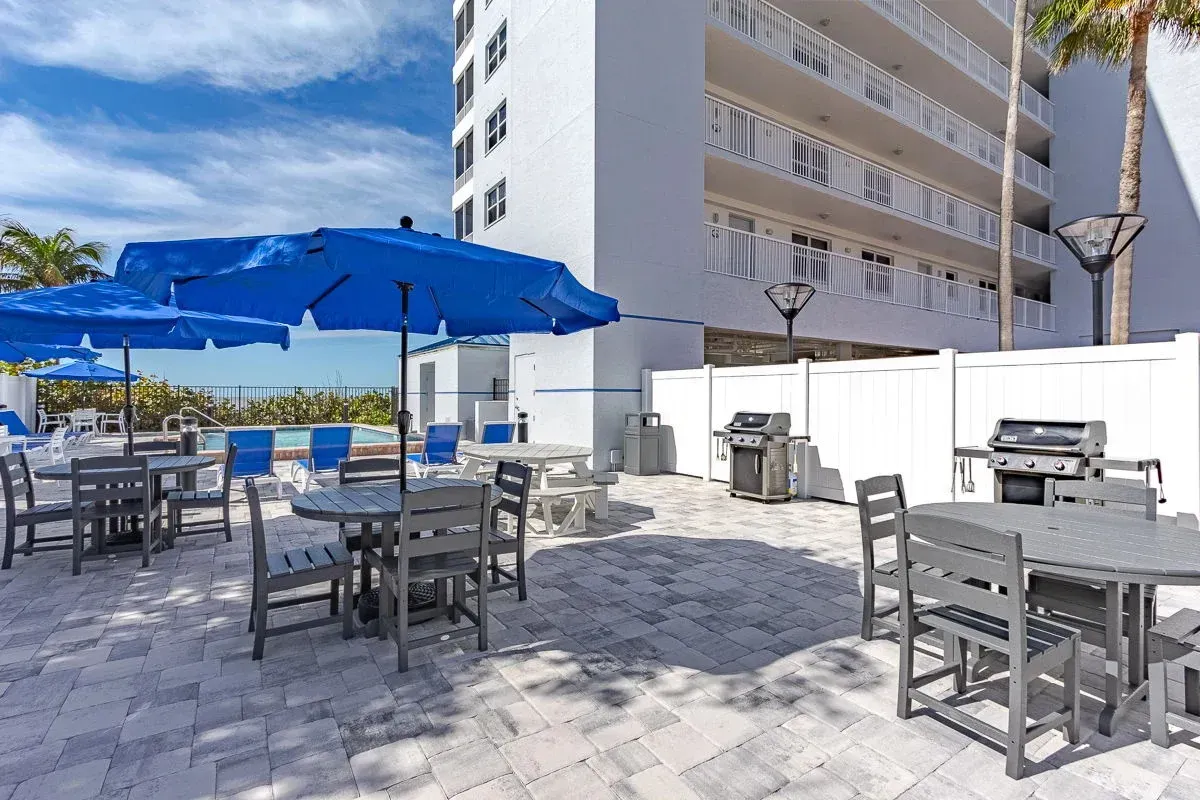 Outdoor patio with tables, chairs, grills, and blue umbrellas, near a swimming pool and building.