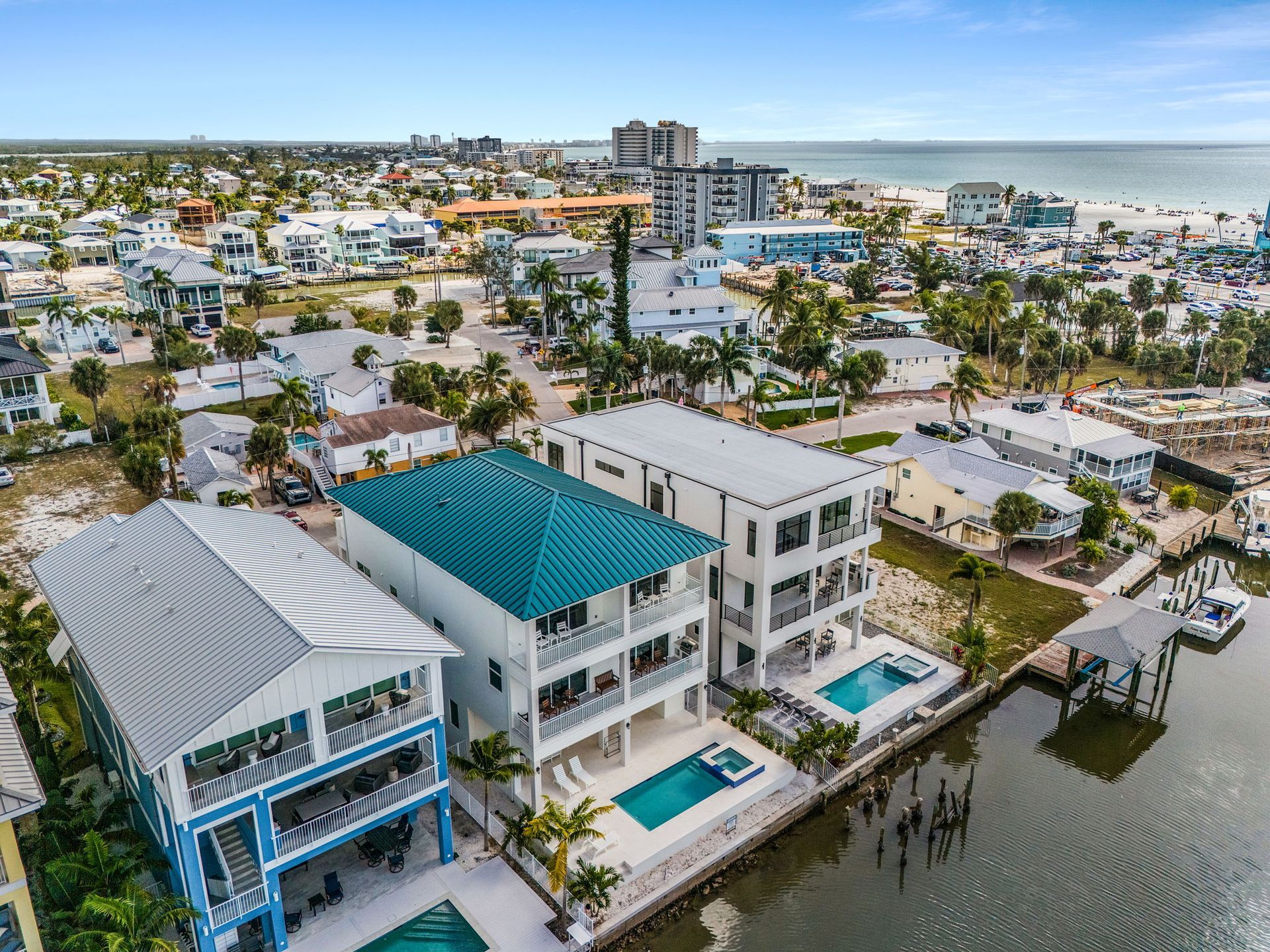 Aerial view of waterfront homes with pools, turquoise roof, and beach in the distance.
