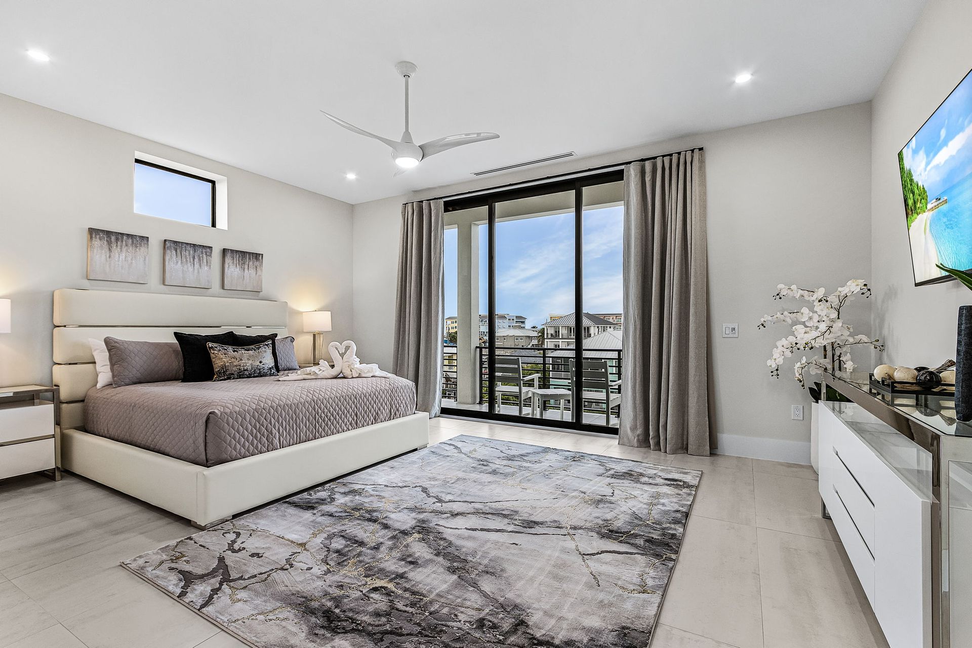 Spacious modern bedroom with bed, rug, balcony, and large TV. Neutral tones with natural light.