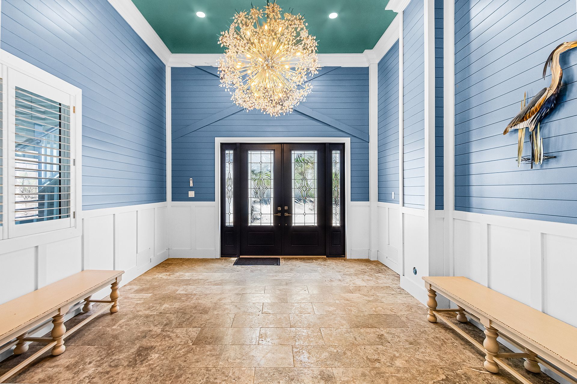 Bright blue hallway with white paneling, chandelier, benches, and black double doors at the end