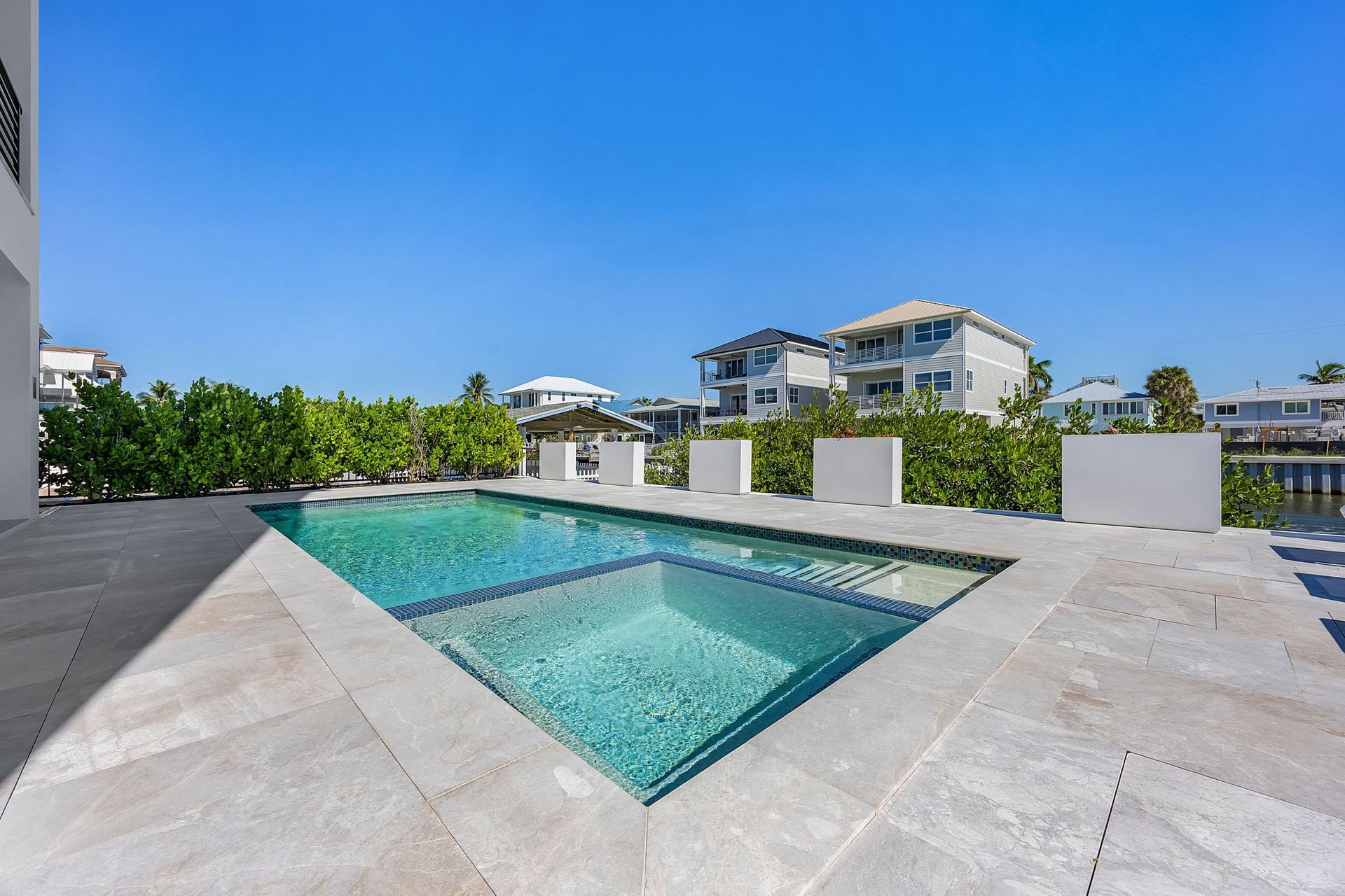 Pool with turquoise water, surrounded by gray stone patio and white planters, with buildings and blue sky in background.