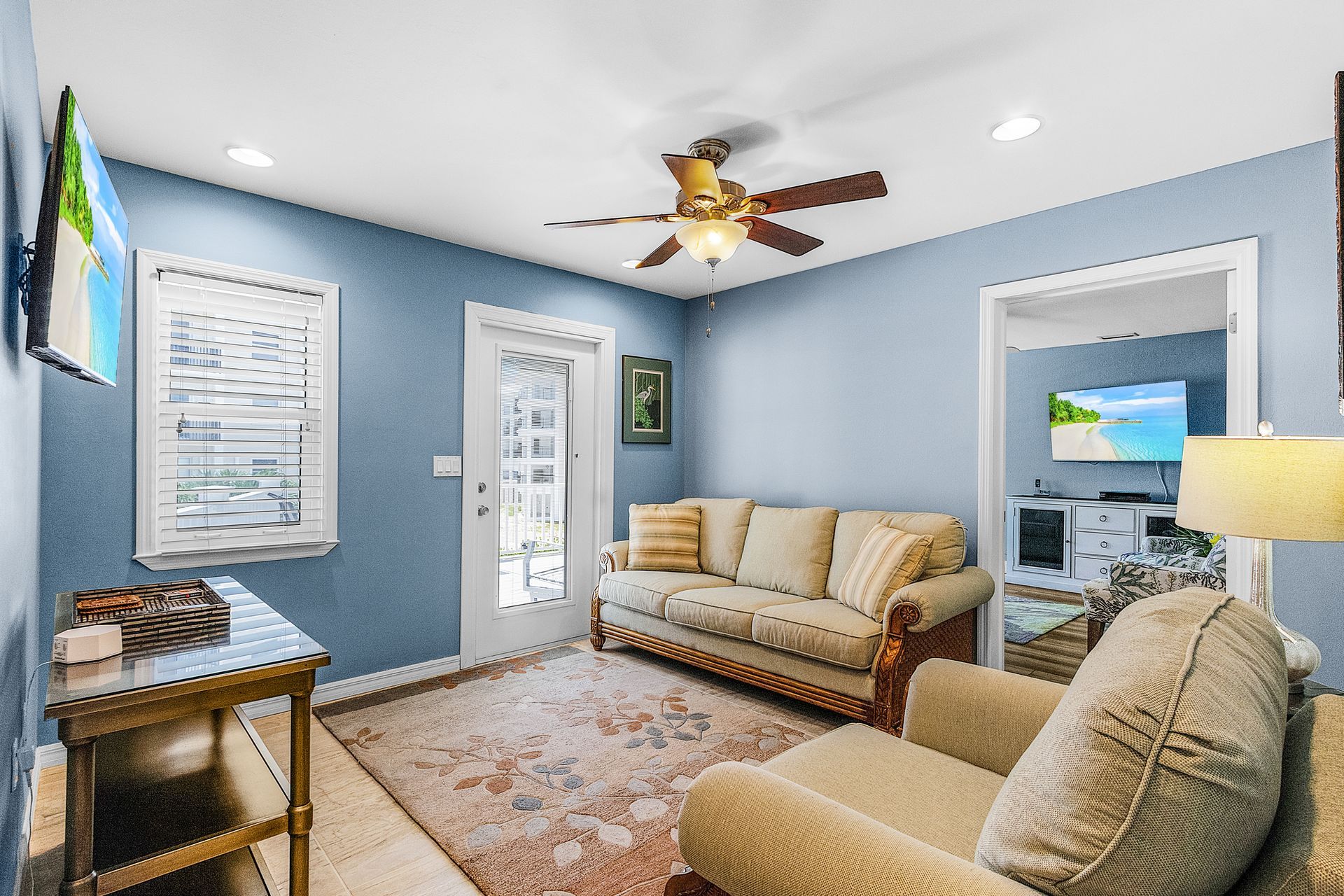 Bright living room with blue walls, beige sofas, patterned rug, ceiling fan, and adjacent doorway to another room