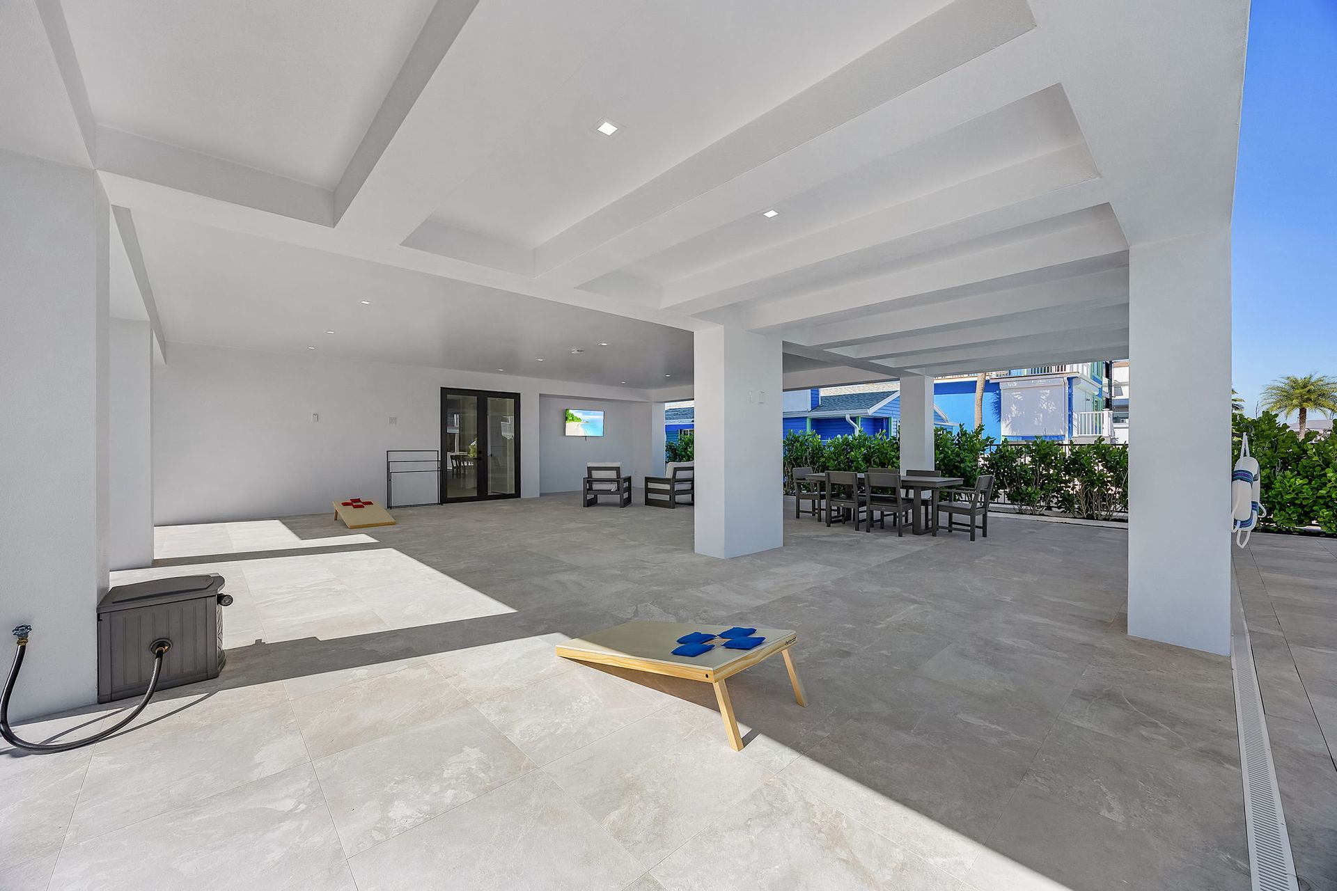 Open-air concrete patio area with a beanbag toss game, tables, and views of a blue building and sky.