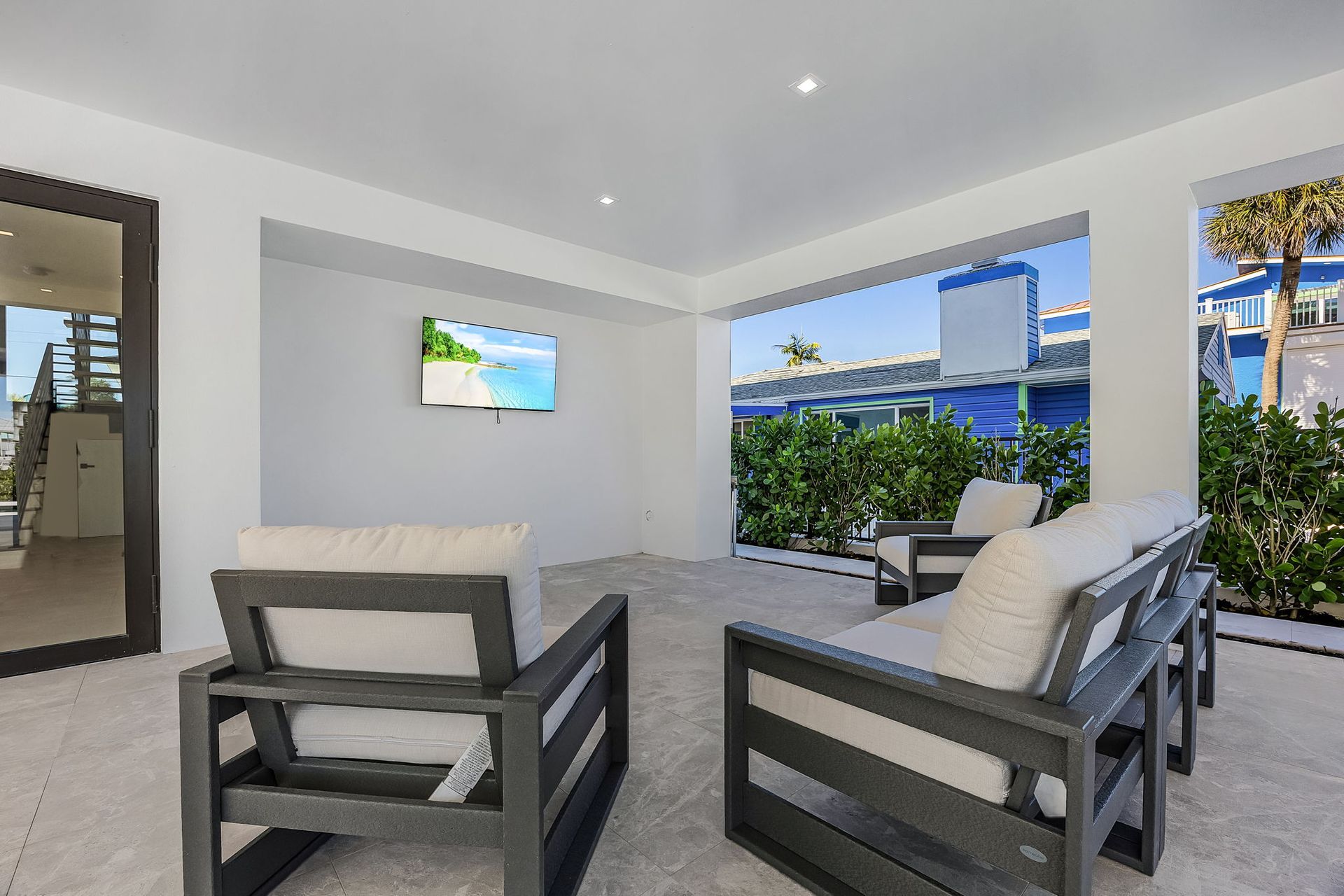 Patio with gray furniture, a TV, and an opening to a view of blue buildings and green foliage.