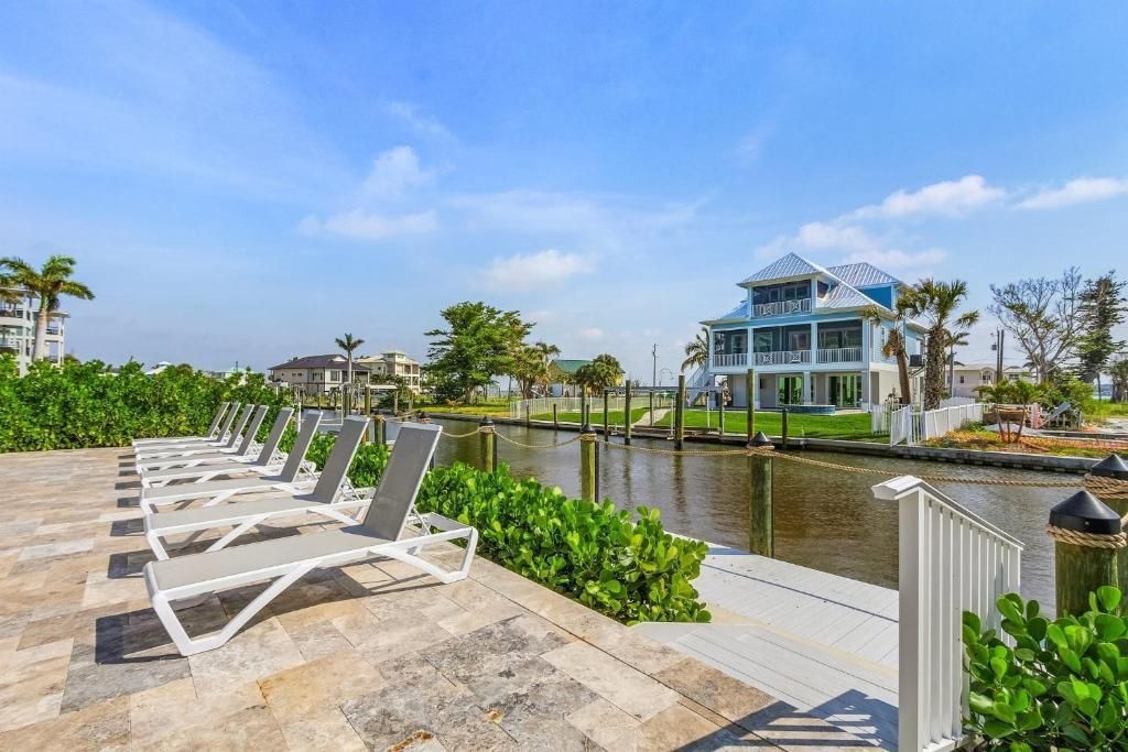 Lounge chairs face a waterway, with a multi-story house visible in the background under a blue sky.