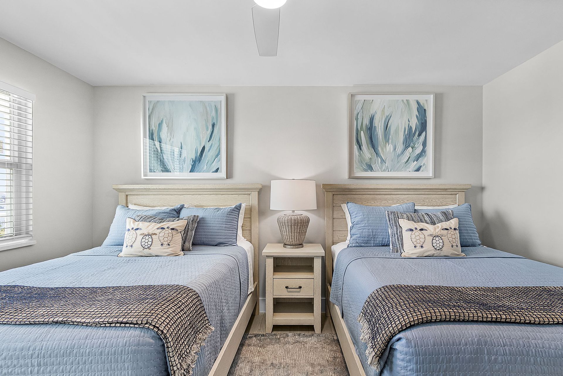 Two twin beds in a bedroom, light blue bedding, nightstand, artwork, white walls, and a window.