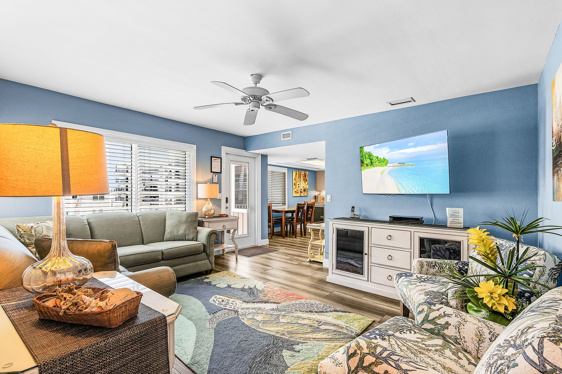 Bright living room with blue walls, gray sofa, white TV console, patterned rug, and large windows with blinds.