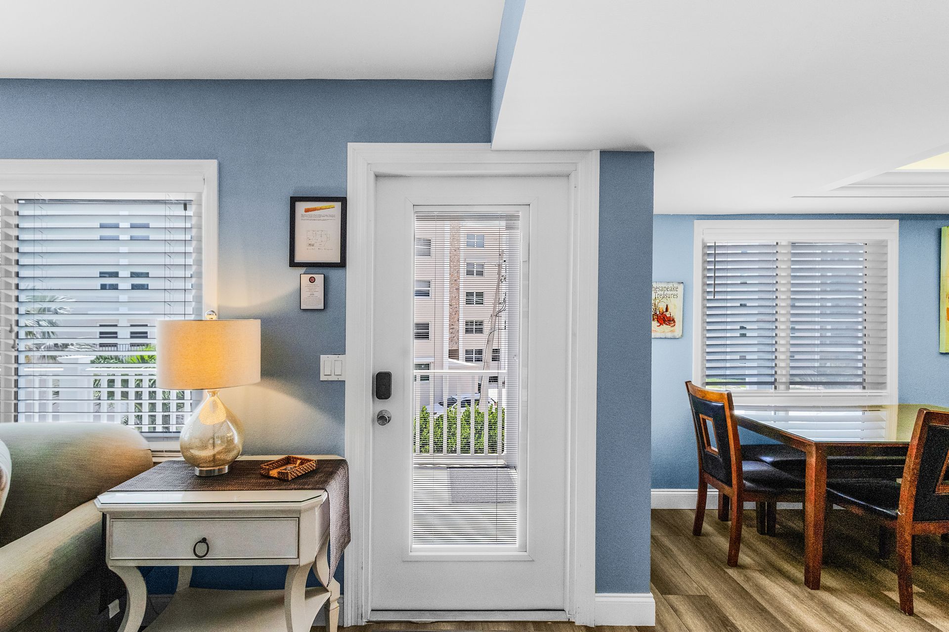 Bright living room with white front door, blue walls, lamp, side table, and dining area by window blinds