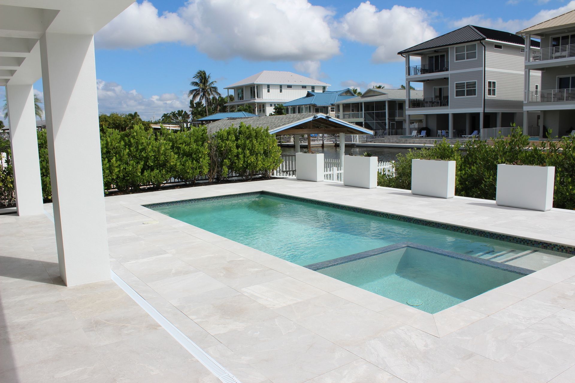 Pool and jacuzzi on a sunny day, surrounded by white structures and greenery; waterfront buildings in the background.