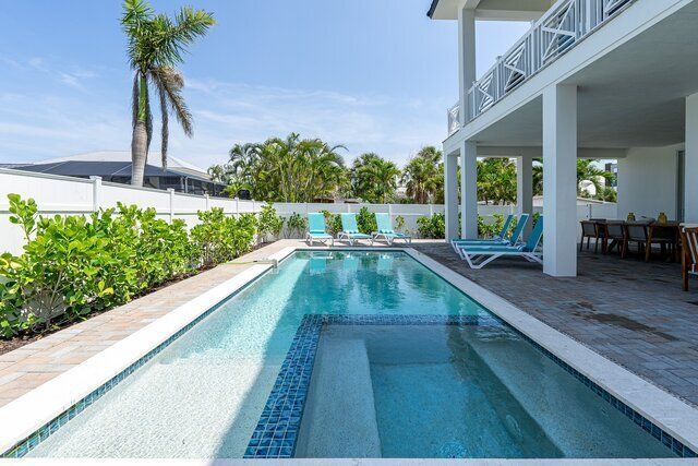 Pool at a white house with palm trees, lounge chairs, and an outdoor dining area.