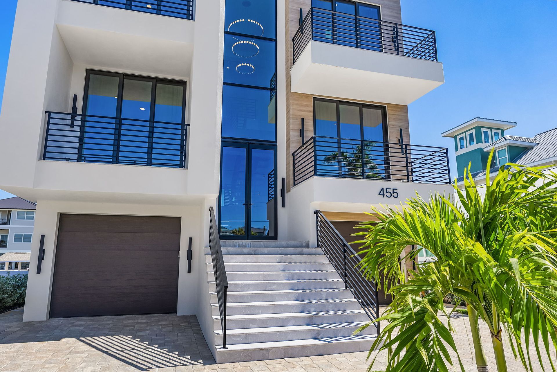 Modern multi-story white house with black railings and large windows, palm tree in front.