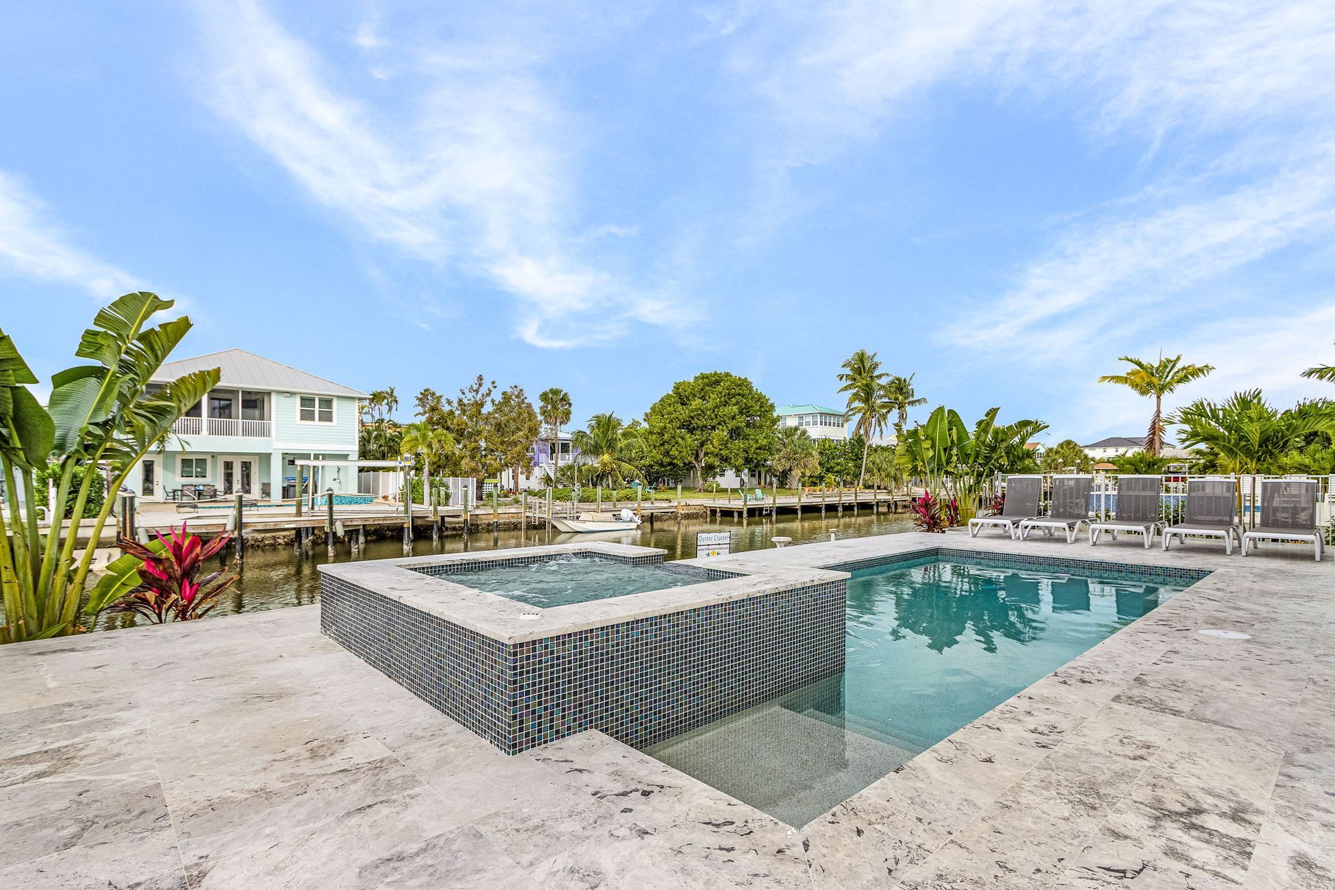 Pool and spa overlooking a canal with houses, palm trees, and lounge chairs; blue sky.