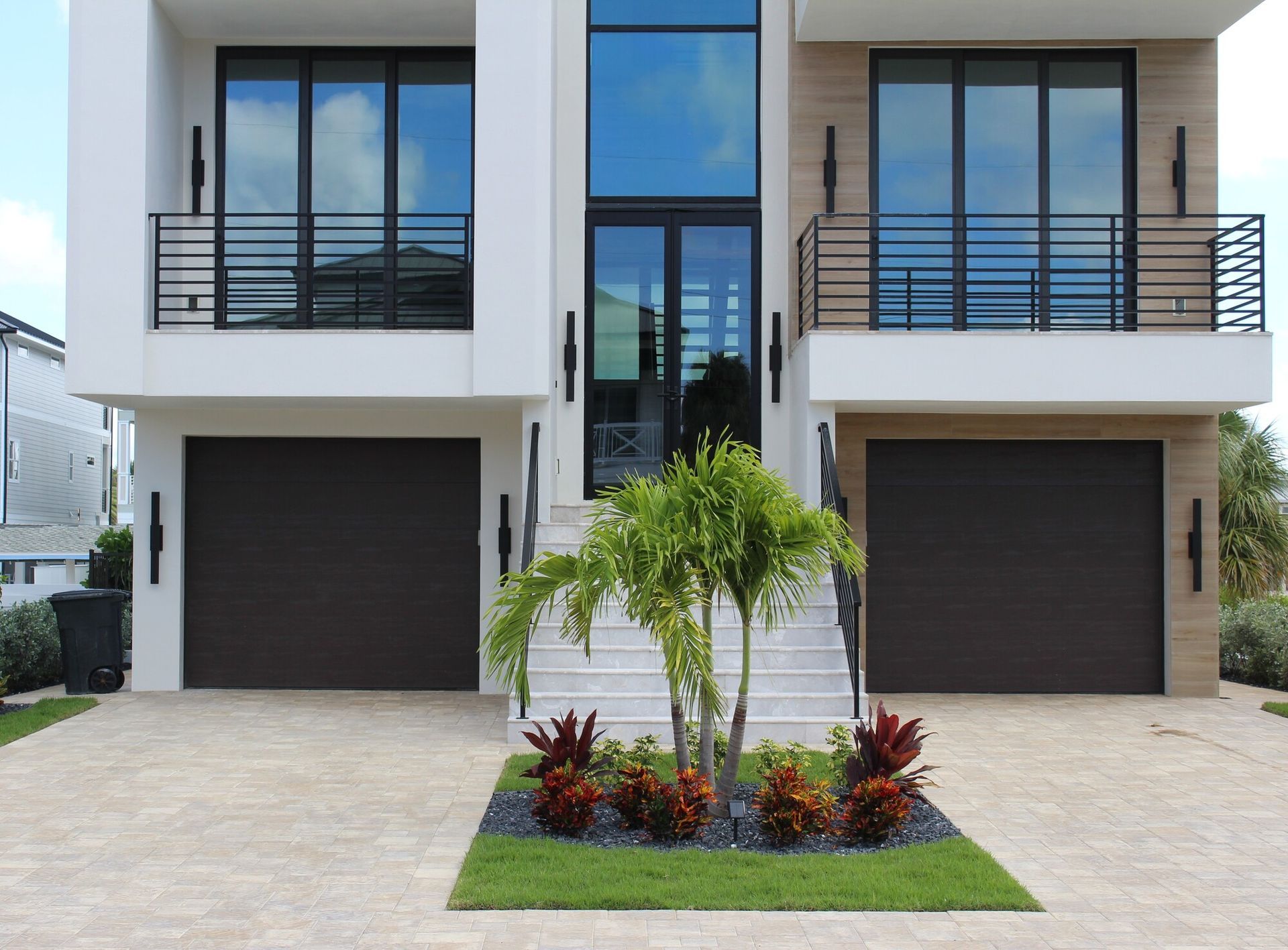 Modern two-story house with dark brown garage doors, balconies, and a central glass door entrance. Landscaping in front.