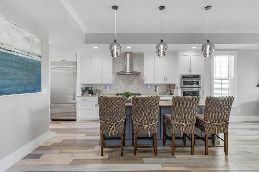 Modern kitchen with island, pendant lights, and upholstered chairs; hardwood floors and white cabinets.