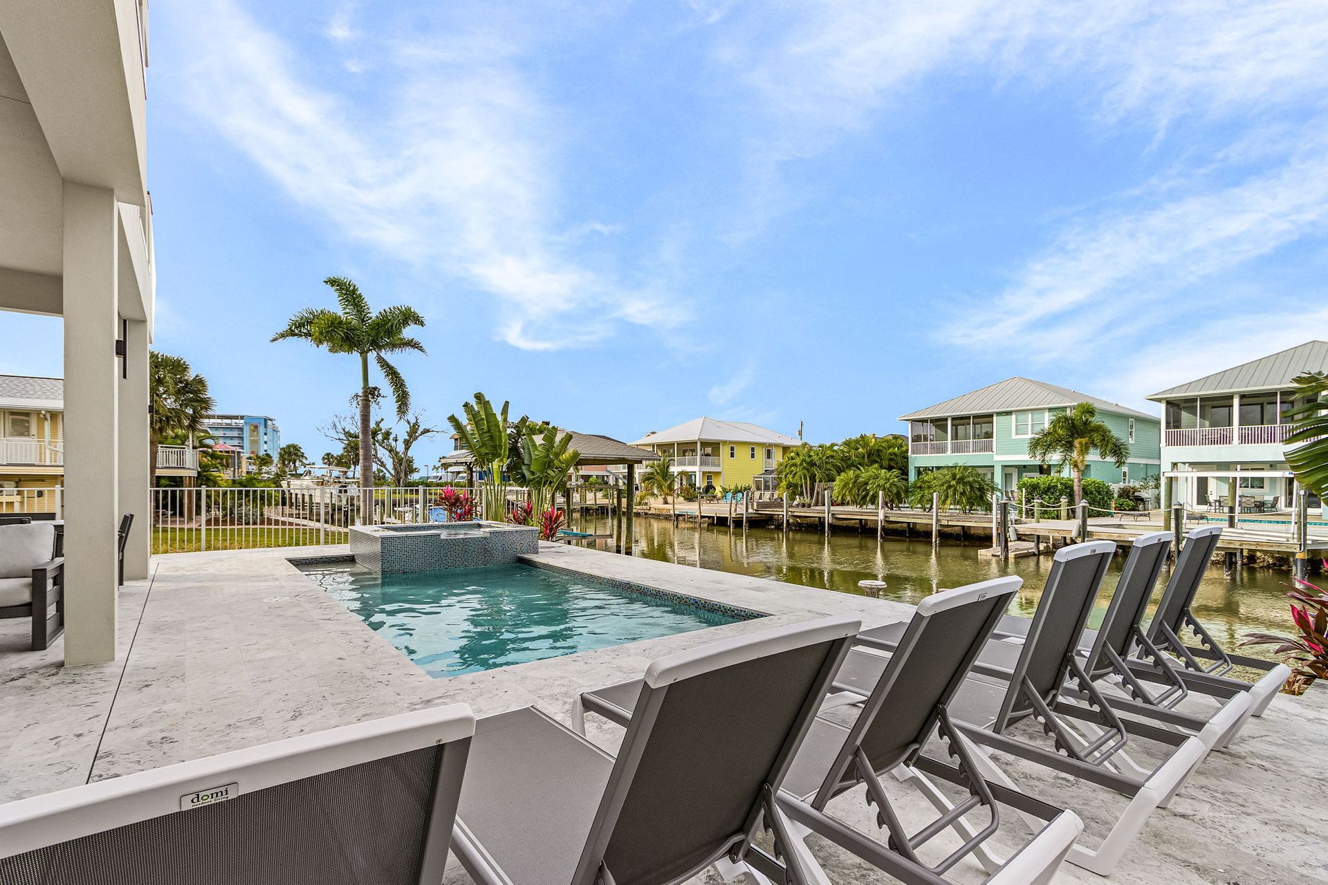 Backyard with pool, canal view, lounge chairs, palm trees, and houses under a cloudy sky.