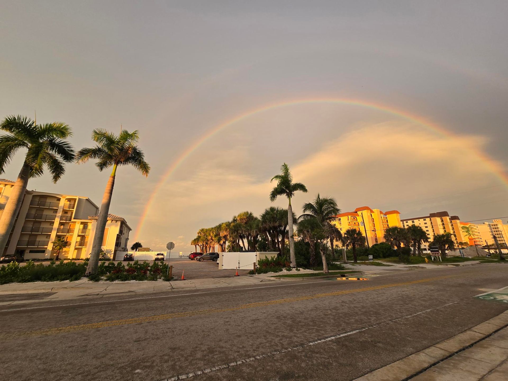 Rainbow arcs over a road lined with palm trees and buildings at sunset.