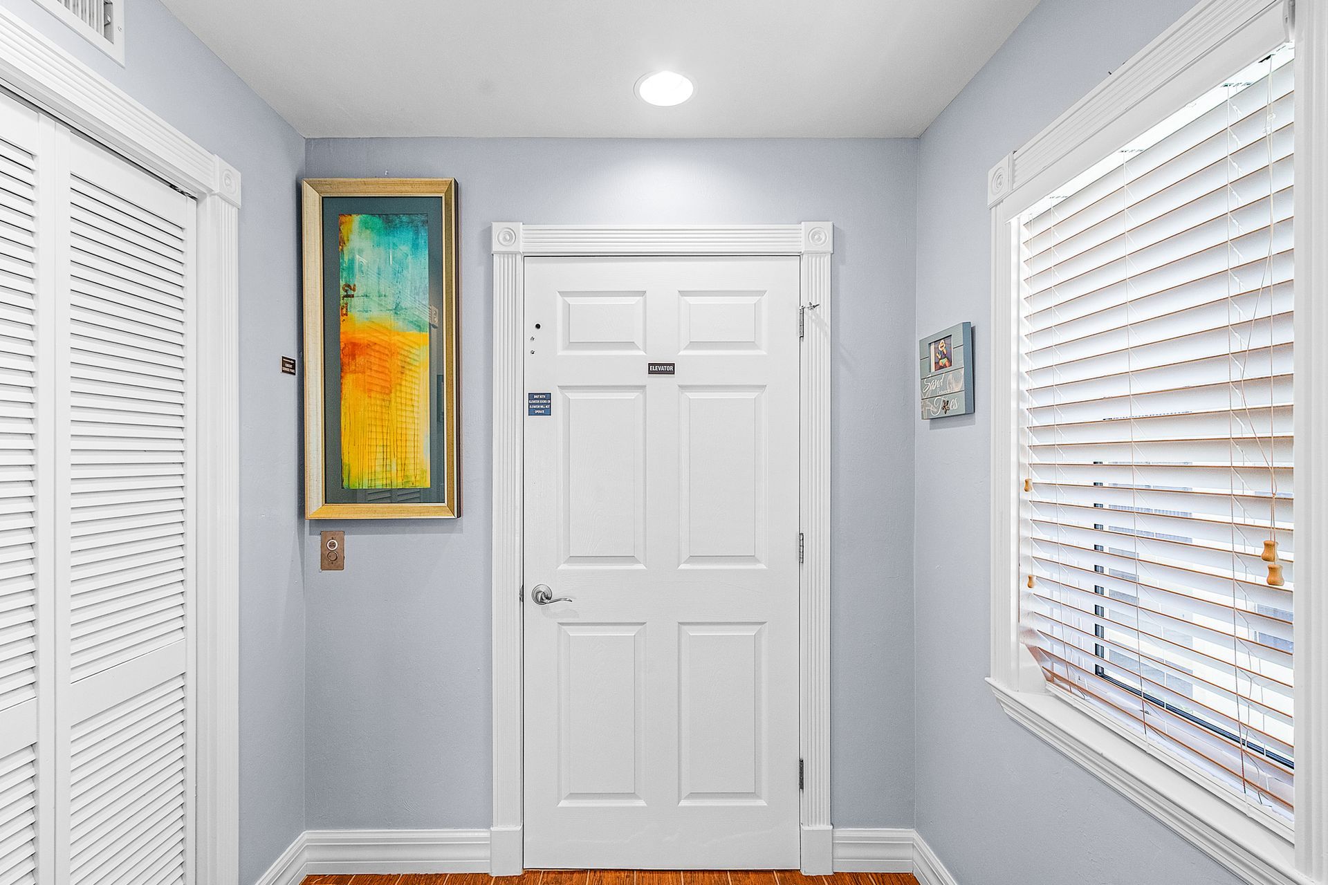 Small white entryway with a white front door, blinds on both sides, and colorful wall art.
