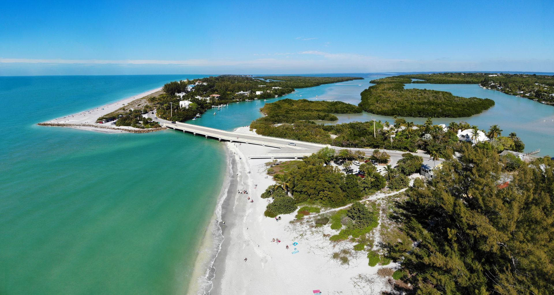 Aerial view: Sandy beach meets turquoise water. Bridge spans waterway lined with green trees under blue sky.