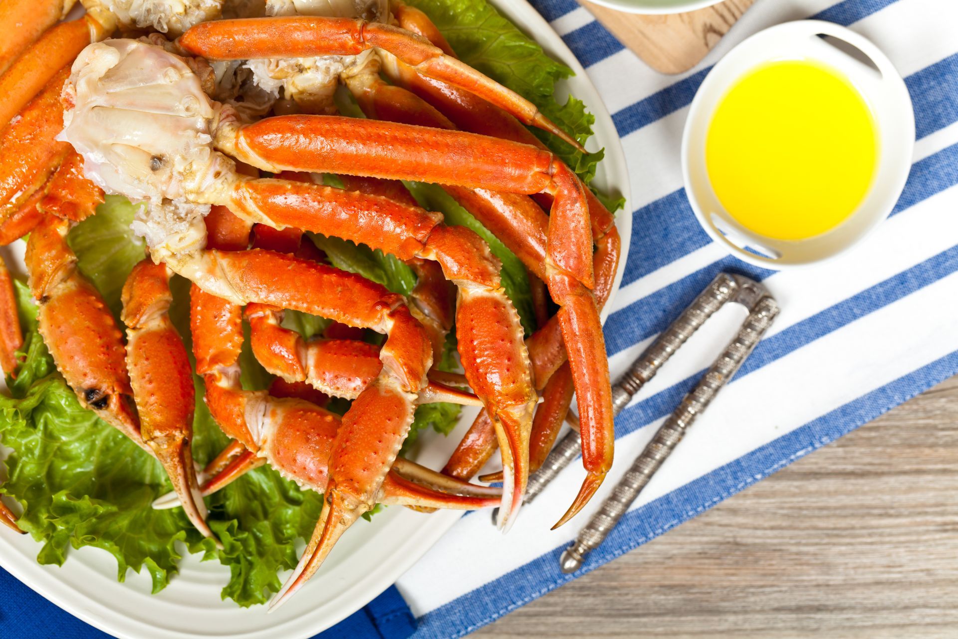Snow crab legs on plate with melted butter, nutcracker, and blue-striped cloth.