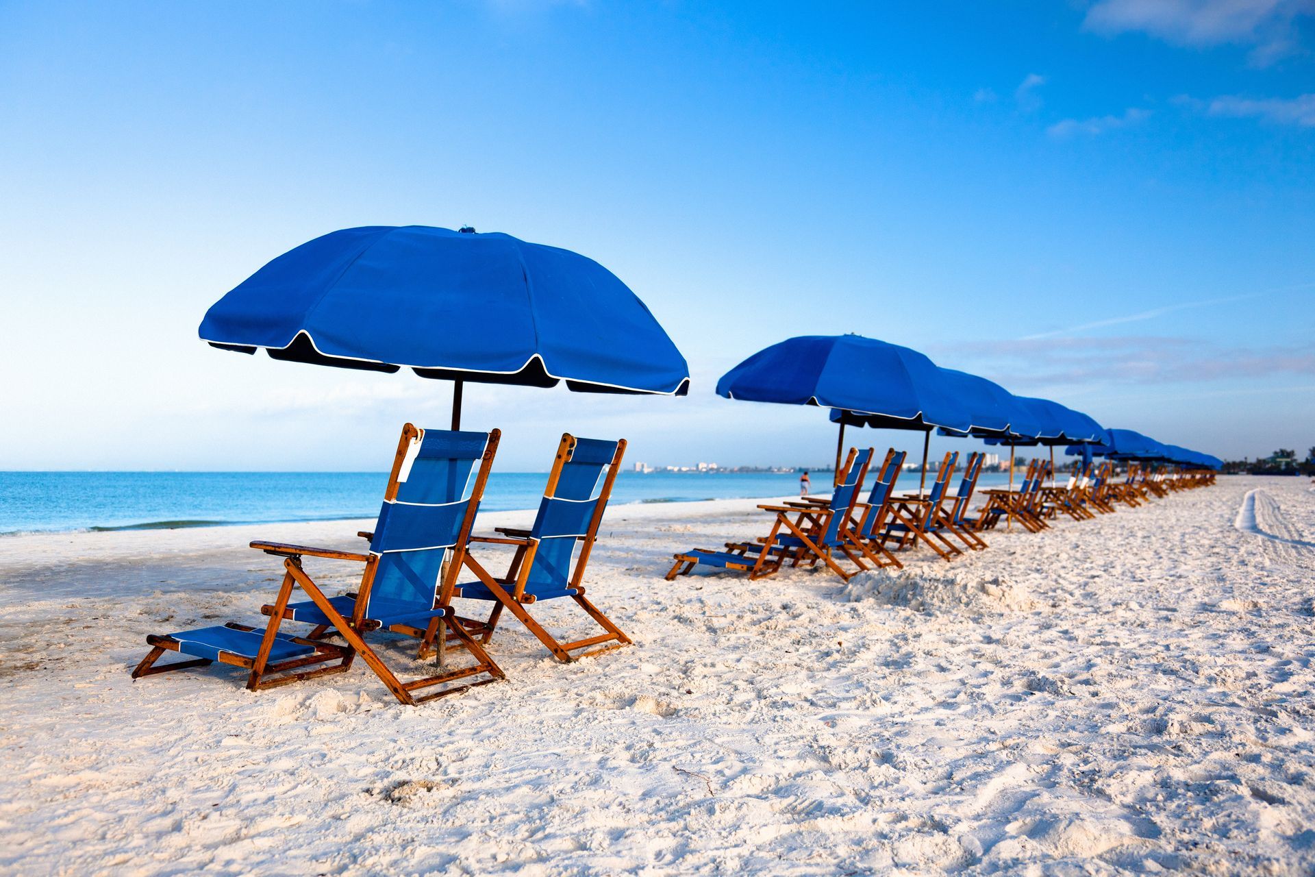 Blue beach umbrellas and chairs line a white sandy beach, overlooking the ocean under a clear blue sky.