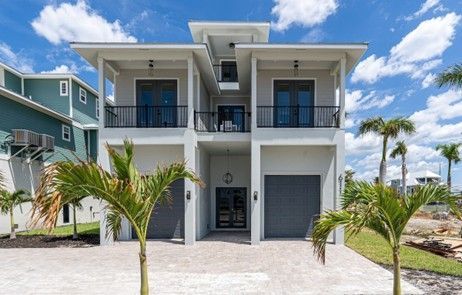 Two-story modern home with gray exterior, balconies, and garage doors. Palm trees and blue sky background.