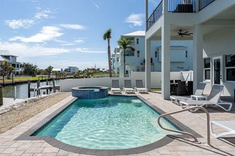Pool and jacuzzi next to a waterfront home with lounge chairs, blue water, and sky.
