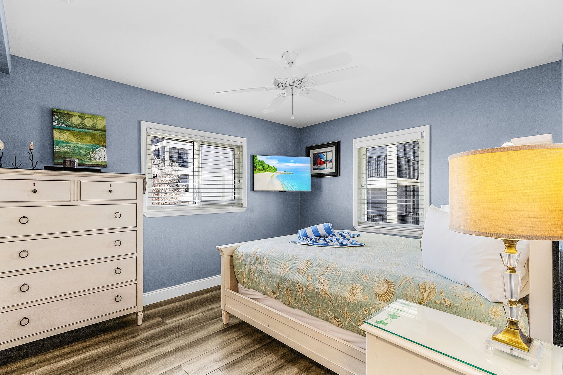 Bright bedroom with blue walls, white dresser, bed, TV, and large windows overlooking greenery