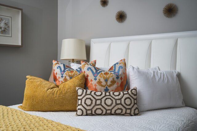 Bed with white headboard, decorative pillows in gold, orange, and patterned textiles, lamp on bedside table.