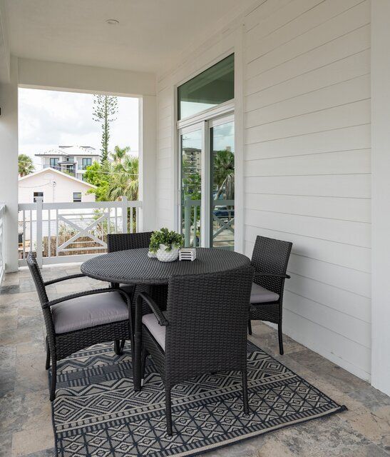 Outdoor dining set on a balcony. Wicker chairs surround a round table on a patterned rug. White walls, window view.