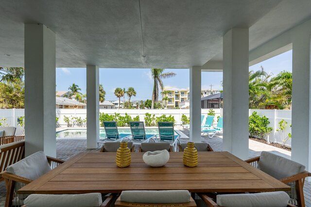 Covered patio with wooden dining table and chairs, overlooking a pool with lounge chairs and palm trees.