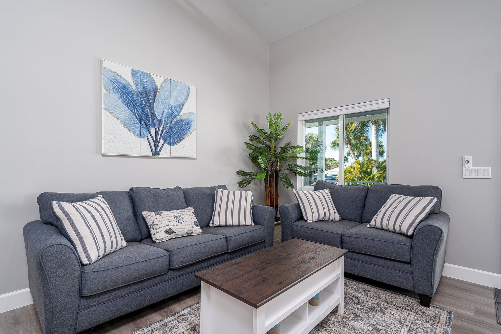 Living room with blue sofas, coffee table, art, and a large plant near a window.