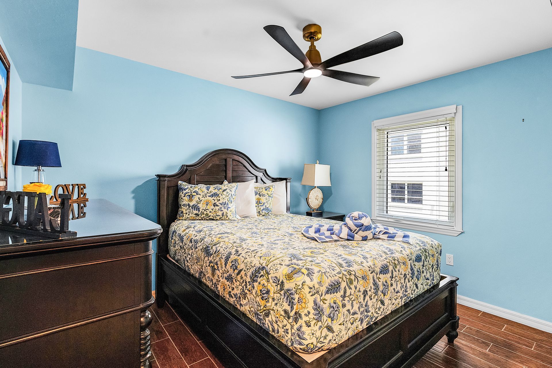 Blue bedroom with dark wood bed, floral bedding, dresser, ceiling fan, and window blinds