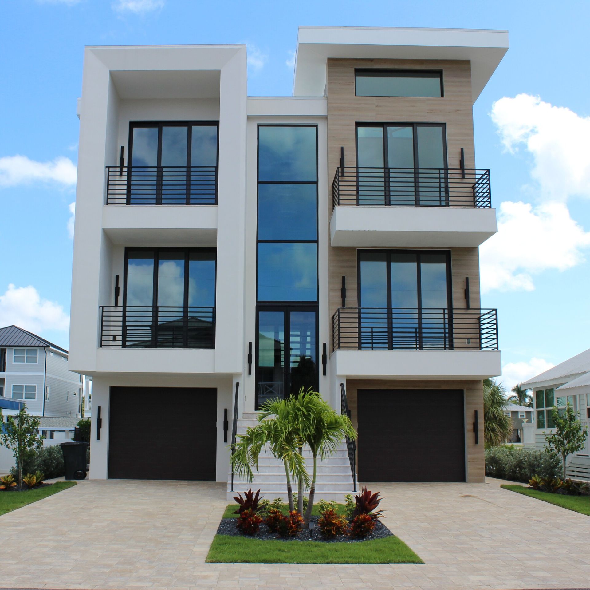 Modern, three-story white house with black framed windows, balconies, and black garage doors, against a blue sky.