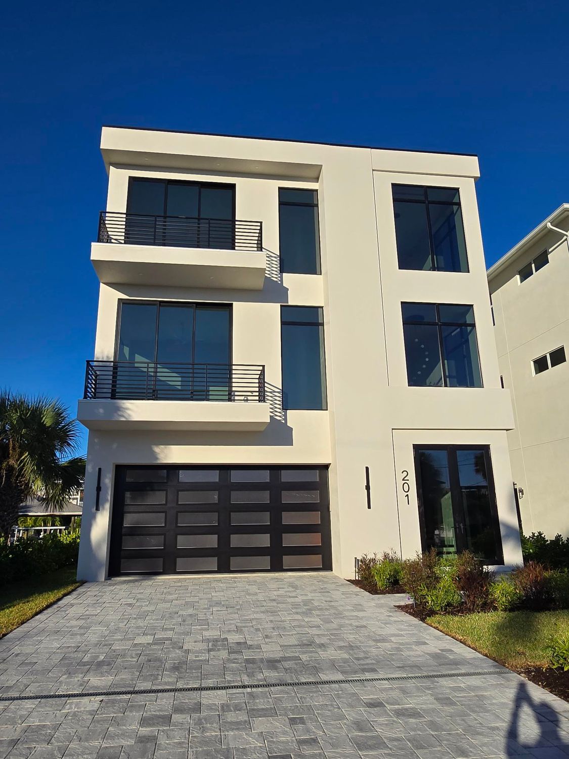 Modern three-story house with a black garage door and gray paved driveway against a clear blue sky.