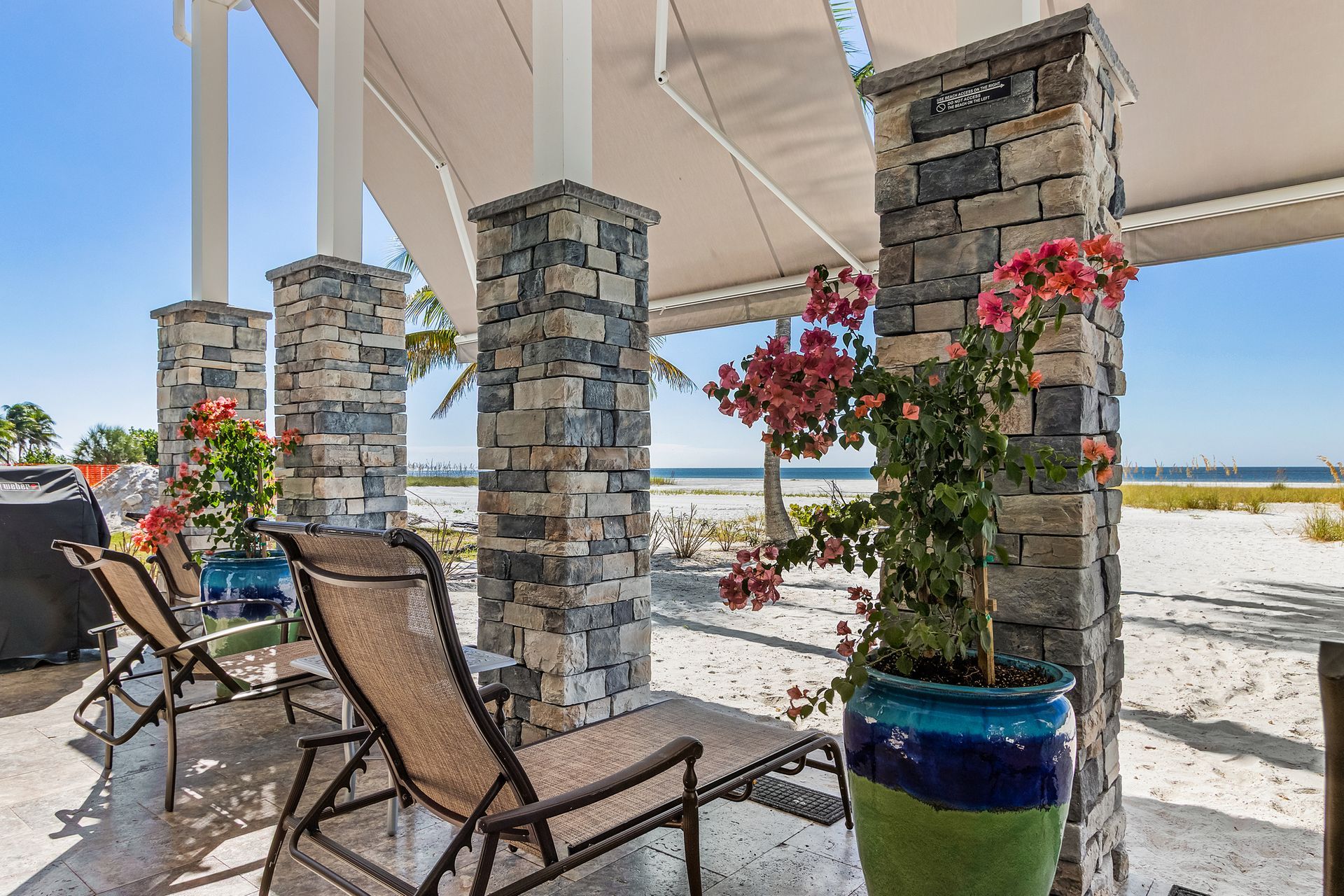 Shaded patio with lounge chairs, stone columns, and potted flowers overlooking a sandy beach and water