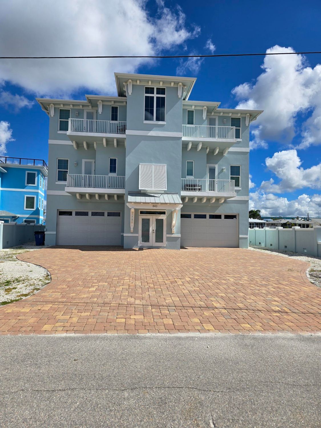 Three-story, light blue beach house with multiple balconies, three-car garage, and brick driveway under a bright sky.