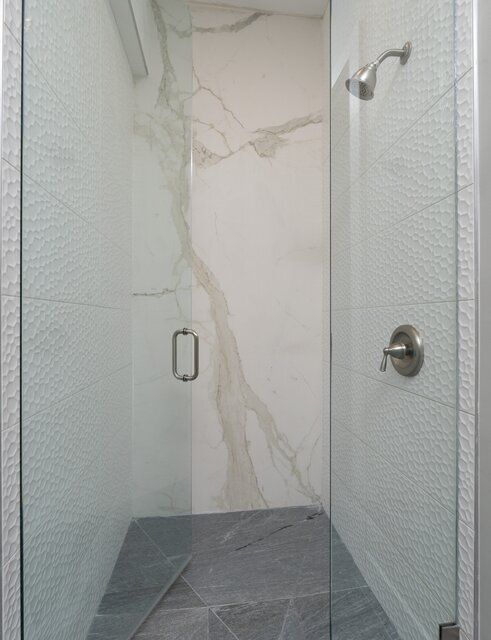 Modern shower stall with glass doors, white and gray tile, marble-like accent wall, and silver fixtures.