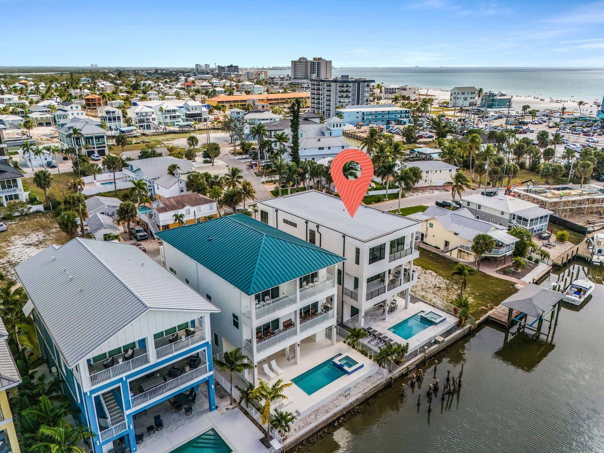 Aerial view of waterfront homes with pools, docks, and ocean views. A red marker pinpoints a white house.