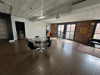 Spacious room with wooden floor, white ceiling, brick wall, round table with black chairs. Windows and black door.