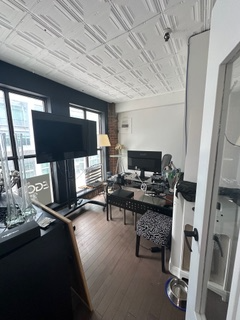 A cluttered home office with a large TV, desk, and zebra-print chair next to a window, leading to an urban view.