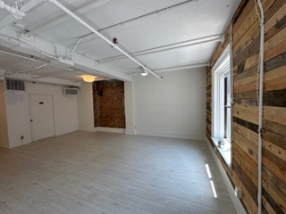 Empty, open-plan room with wood-paneled wall, white walls and ceiling, and a large window. The floor is light-colored.