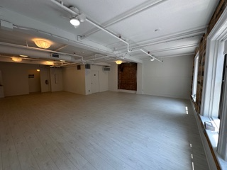 Empty, bright white room with tall windows and exposed pipes. Some brick wall sections are visible.