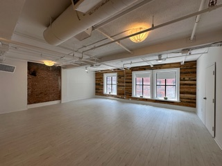 Empty studio space with white walls, white floor, brick and wood accents, and two windows.