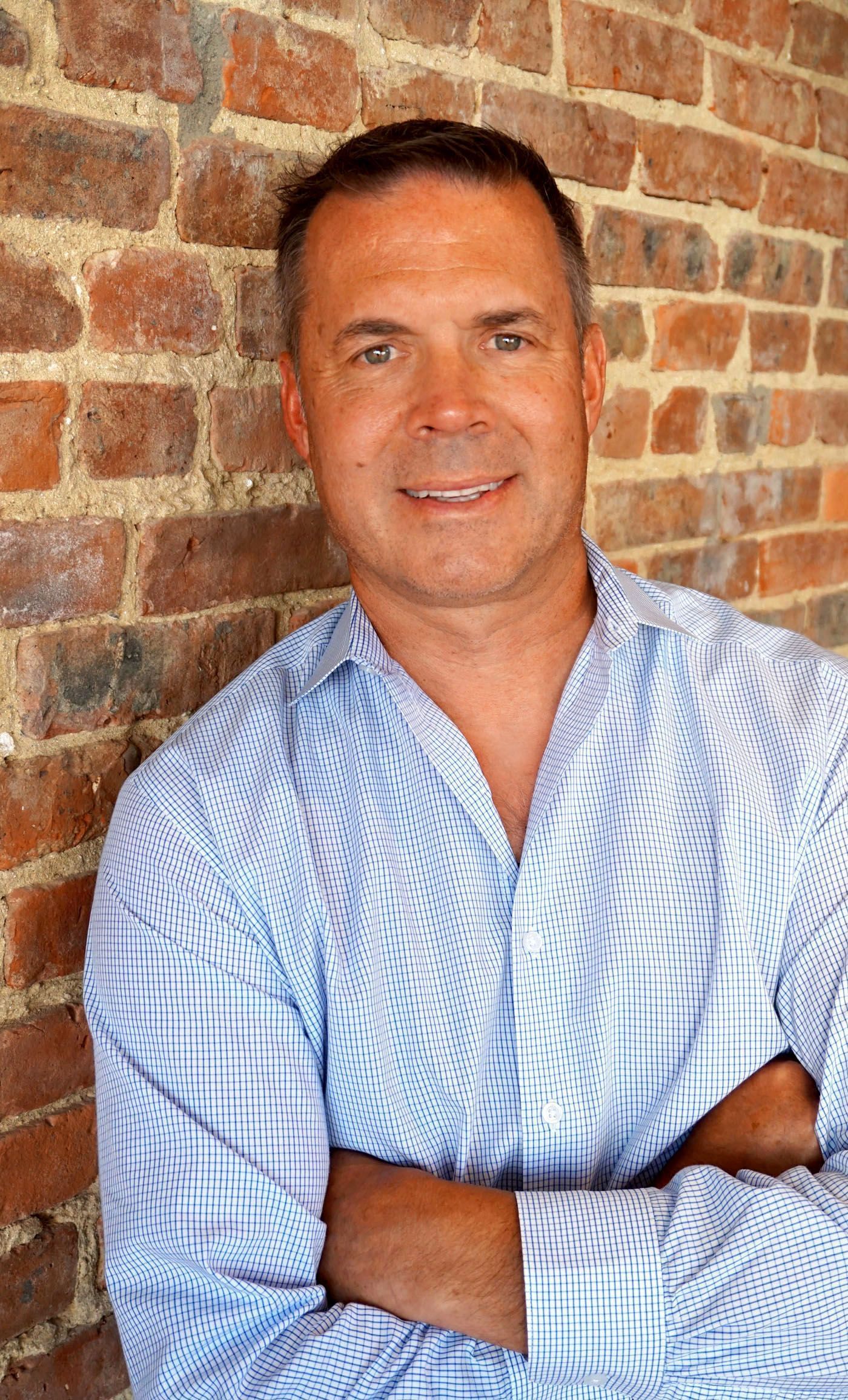 Man in blue patterned shirt leans against brick wall, arms crossed, smiling.