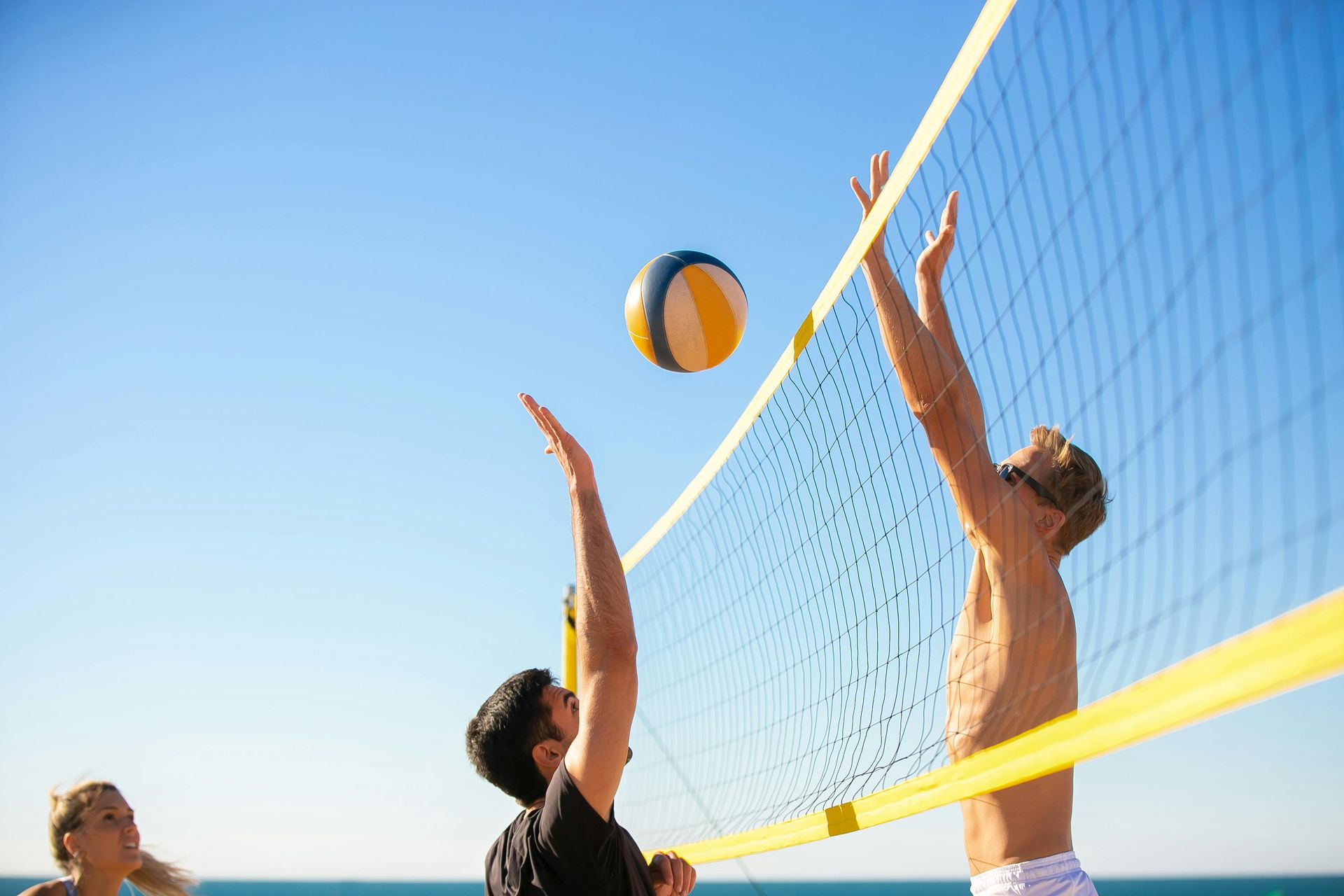 Beach volleyball: Players near net, reaching for ball in mid-air on sunny day.