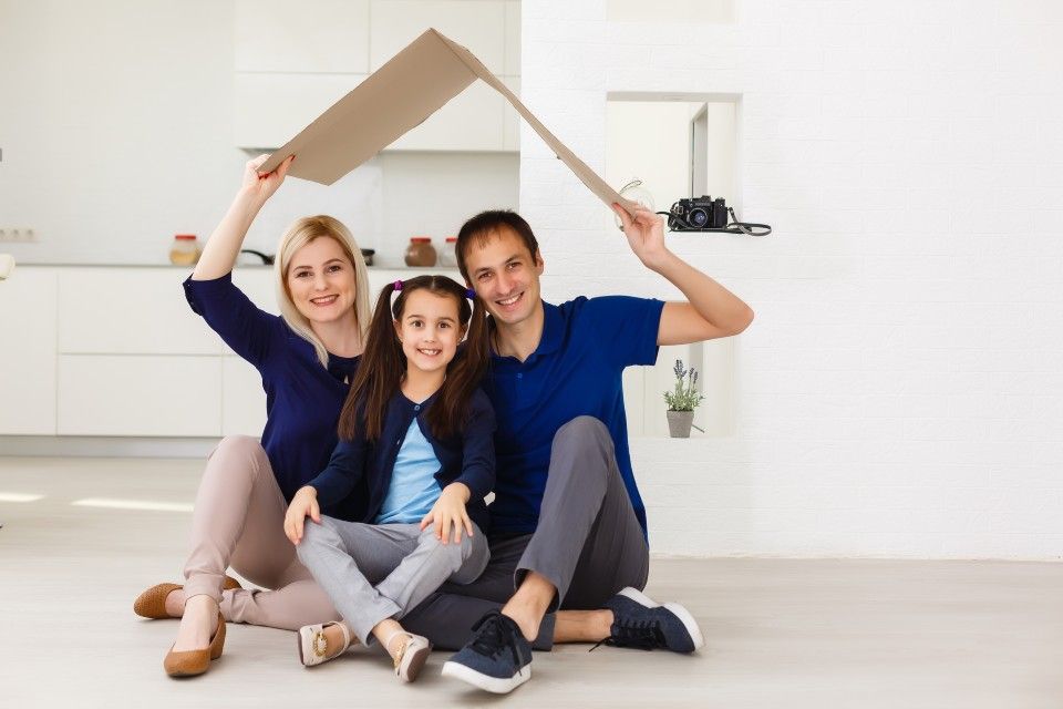 Family of three sitting, holding a cardboard house over their heads, smiling in a bright room.