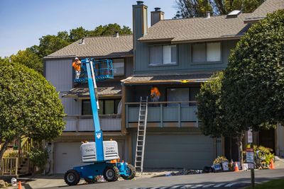 Construction workers on a blue boom lift renovating a multi-story building with a gray facade and brown roof.