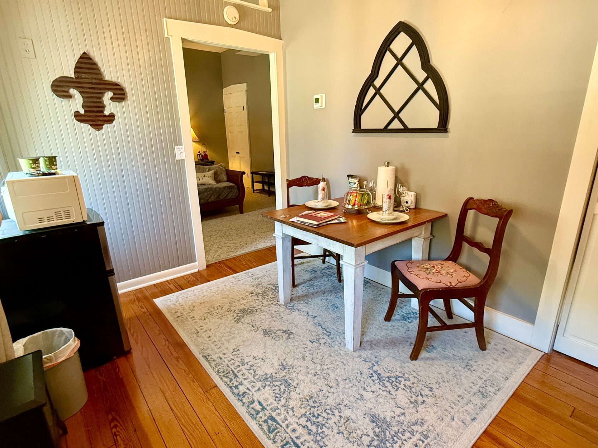 A small dining area with a table set for two, rug on wooden floors, and a glimpse into another room.